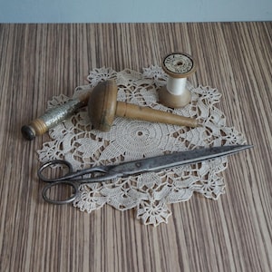 Op de afbeelding: Een verzameling naaigerei bestaande uit een vintage metalen schaar, een houten stop-ei en een kleine houten klos met wit garen, rustend op een rond, crèmekleurig gehaakt kanten kleedje met een bloemmotief.