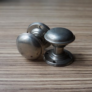 Op de afbeelding: Drie antieke zilverkleurige deurknoppen met een afgerond ontwerp. De knoppen hebben een cirkelvormige basis met schroefgaten en een gewelfde handgreep. De hardware is geplaatst op een lichtbruin houten oppervlak.