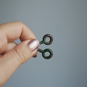 Könnte beinhalten: Zwei kleine, vintage Metall-Augenbolzen mit grüner Oberfläche und einem kreisförmigen Auge. Die Augenbolzen werden vor einem grauen Hintergrund gehalten. Das Metall hat ein abgenutztes, antikes Aussehen.