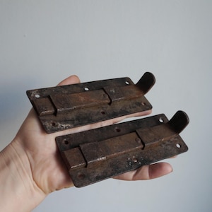 May include: Two antique, rectangular, rusty metal slide bolts. Each bolt has a sliding bar and a curved handle. The bolts have several holes for mounting. The bolts are held in a hand.