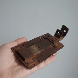 Puede incluir: Un pestillo metálico oxidado, de forma rectangular, con un cerrojo corredero y dos soportes angulados. El pestillo tiene múltiples orificios para tornillos y muestra signos de desgaste. El metal tiene un aspecto marrón y envejecido.