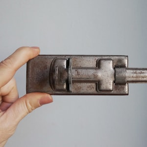 Pode incluir: Um ferrolho de metal retangular e antigo com um mecanismo deslizante. O ferrolho é de cor prateada opaca e envelhecida, com sinais de desgaste. O ferrolho é segurado em uma mão contra um fundo simples. Este item é provavelmente usado para proteger portas ou portões.