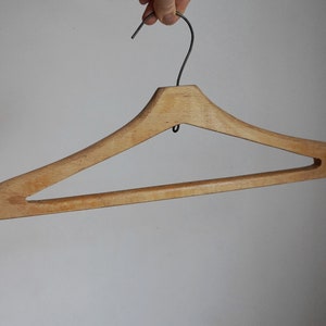 May include: A wooden clothes hanger with a metal hook. The hanger is made of light-colored wood and has a classic design. The hook is a dark gray color. The hanger is designed for hanging clothing.