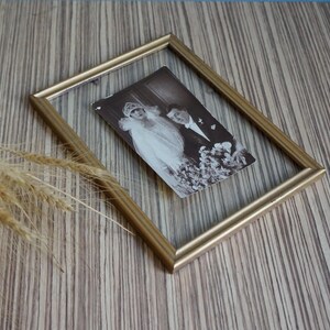 May include: A rectangular gold-toned floating picture frame containing a black and white vintage photograph of a person in a wedding veil and a person in a suit. The frame has a thin metallic border and clear glass panels on both sides.