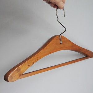 Peut inclure: Un cintre en bois de couleur marron clair, avec un crochet en métal argenté. Le cintre est tenu par une main sur un fond blanc uni. Le design est incurvé.