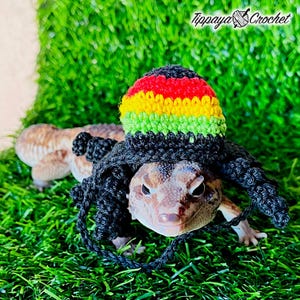 May include: A leopard gecko wearing a crocheted hat in the colors of red, yellow, green, and black, with matching dreadlocks. The gecko is on a bed of green artificial grass. The text "Tippaya Crochet" is in the upper right corner.