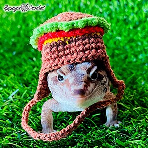 May include: A gecko wearing a crocheted hat designed to look like a hamburger. The hat features green, red, yellow, and brown yarn. The gecko is on a bed of green artificial grass. The text "Tippaya Crochet" is in the upper left corner.