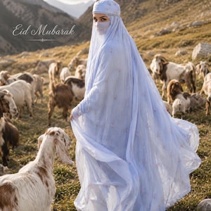 May include: A light blue abaya with a matching head covering and face veil, featuring delicate floral embroidery throughout the fabric. The garment is long and flowing, with the text Eid Mubarak visible in the upper left corner of the image.