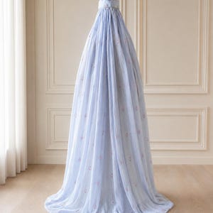 May include: A light blue, floor-length, conical fabric canopy featuring a delicate, repeating pattern of small pink floral motifs. The structure is gathered at the top with a circular cap and drapes downward in soft, vertical folds to the floor.