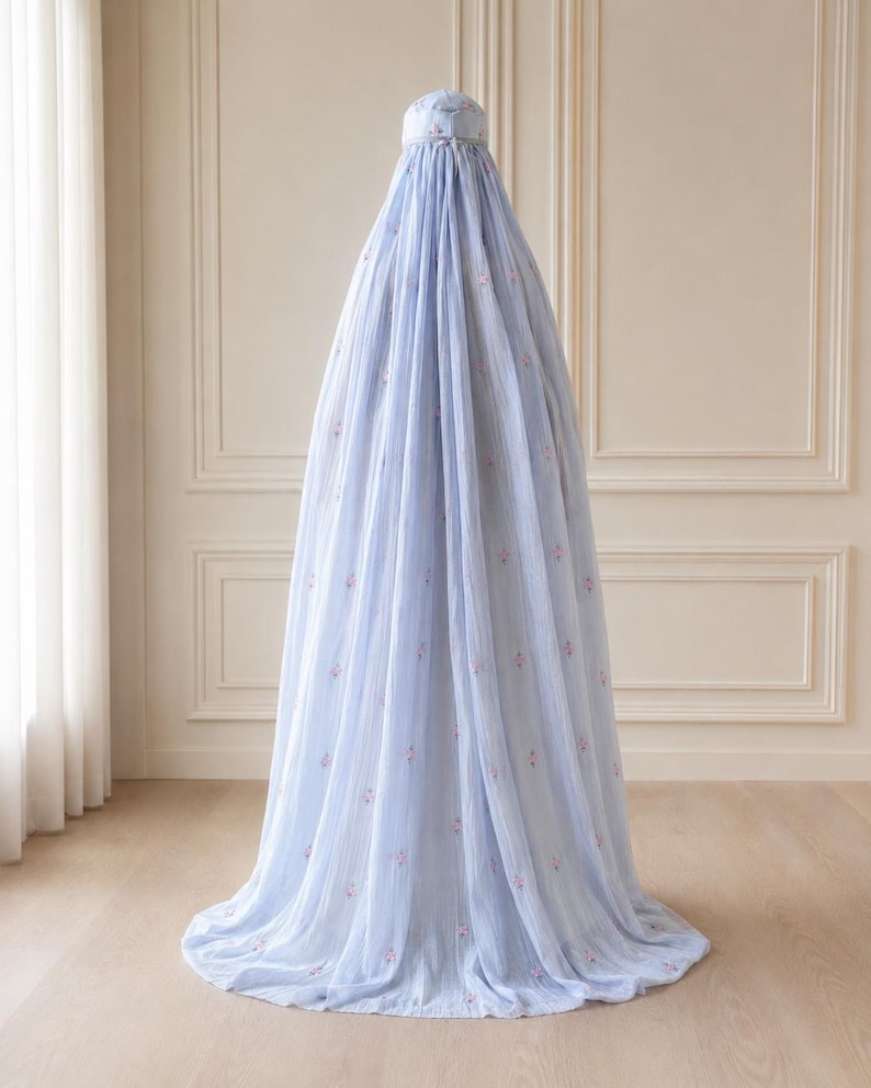 May include: A light blue, floor-length, conical fabric canopy featuring a delicate, repeating pattern of small pink floral motifs. The structure is gathered at the top with a circular cap and drapes downward in soft, vertical folds to the floor.