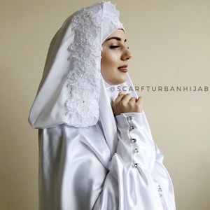 May include: White satin hijab with a lace trim and pearl embellishments. The hijab has a hood and long sleeves with button closures. The text "@scarfturbanhijab" is visible in the image.