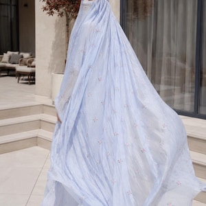 May include: A light blue, floor-length abaya featuring a delicate, repeating floral pattern in soft pink and white. The garment includes a matching headpiece and a face veil, crafted from a lightweight, flowing fabric that drapes elegantly.