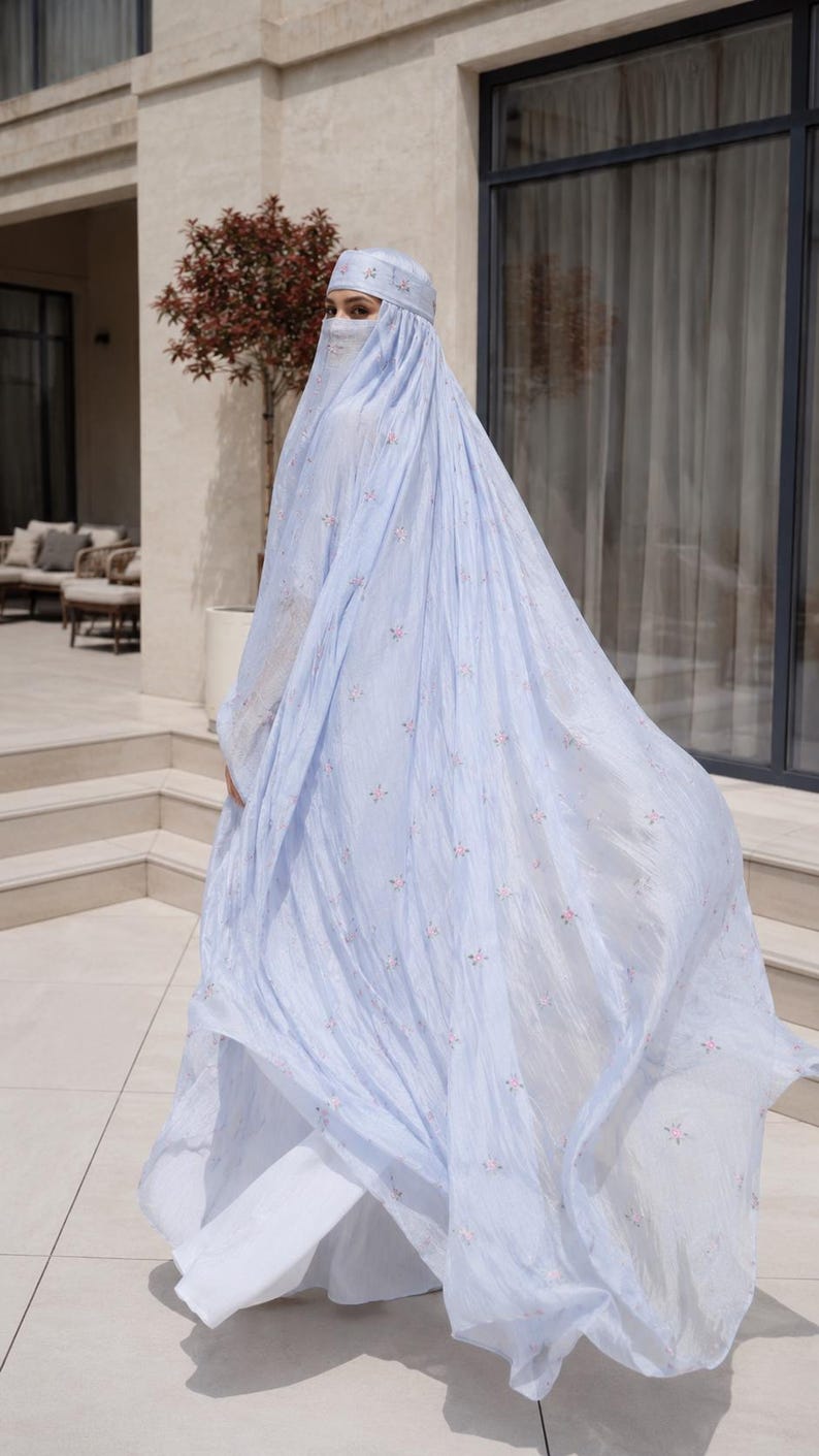 May include: A light blue, floor-length abaya featuring a delicate, repeating floral pattern in soft pink and white. The garment includes a matching headpiece and a face veil, crafted from a lightweight, flowing fabric that drapes elegantly.
