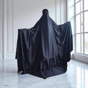 Può includere: Un niqab nero, un indumento indossato da alcune donne musulmane, è esposto in una stanza luminosa. Il niqab copre l'intero corpo, con un'apertura a rete per gli occhi. Il tessuto drappeggia elegantemente, creando una silhouette fluida.