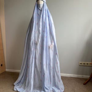 May include: A light blue, floor-length prayer garment featuring a matching head covering and a face veil. The fabric is adorned with a delicate, repeating pattern of small pink floral embroidery throughout the entire length of the flowing material.