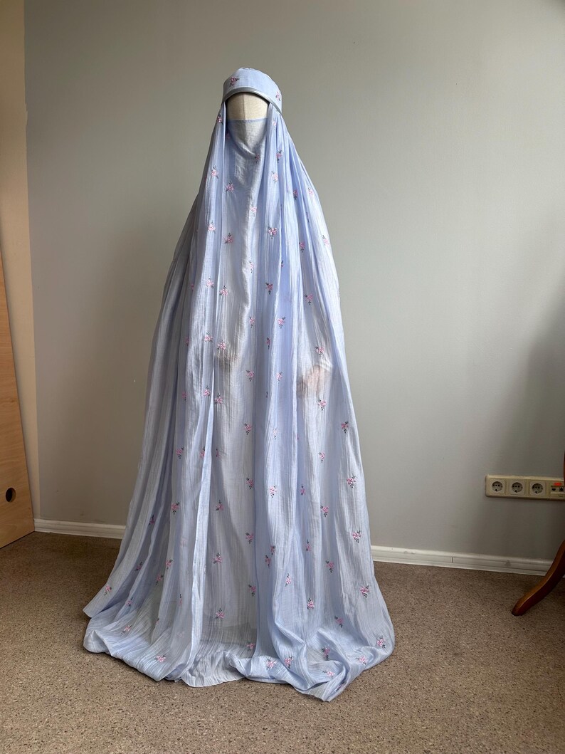 May include: A light blue, floor-length prayer garment featuring a matching head covering and a face veil. The fabric is adorned with a delicate, repeating pattern of small pink floral embroidery throughout the entire length of the flowing material.