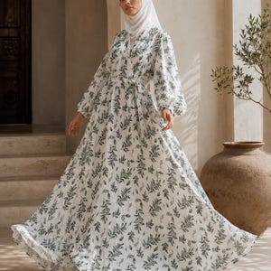 Op de afbeelding: Een witte maxi-jurk met een groen bladerpatroon. De jurk heeft lange, volumineuze mouwen, een wikkelbustier en een bijpassende riem. Het model draagt een witte hijab. De jurk is geschikt voor bescheiden kleding.