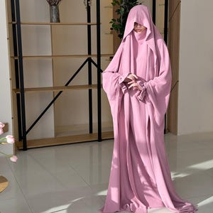 May include: A long, pink, hooded robe with long sleeves. The robe is made of a soft, flowing fabric.