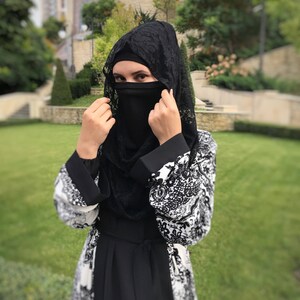 May include: A woman wearing a black abaya with a white floral pattern and a black lace hijab. She is adjusting the hijab, which covers her face.