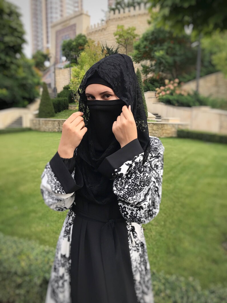 May include: A woman wearing a black abaya with a white floral pattern and a black lace hijab. She is adjusting the hijab, which covers her face.