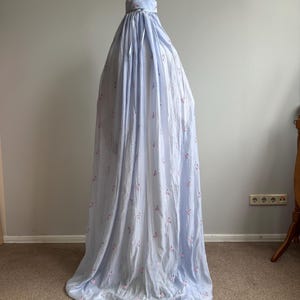 May include: A light blue, floor-length prayer garment featuring a delicate pattern of small pink floral sprigs. The piece includes a fitted head covering section with a matching ribbon tie at the neck, flowing down into a loose, pleated silhouette.
