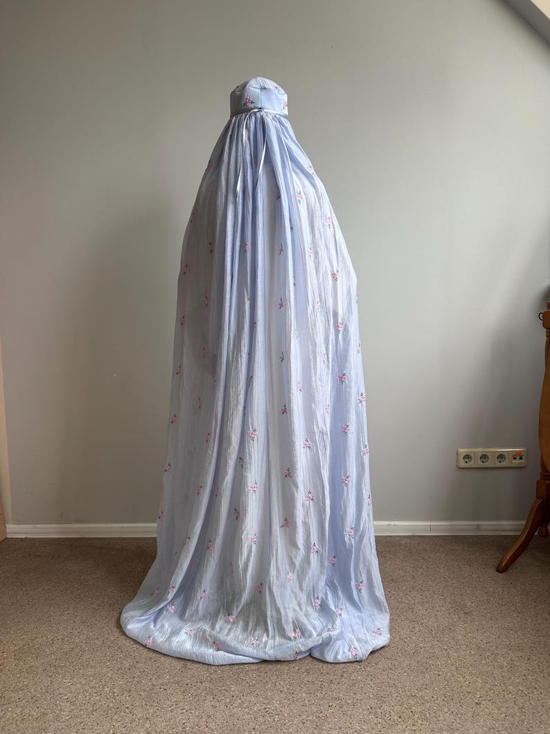 May include: A light blue, floor-length prayer garment featuring a delicate pattern of small pink floral sprigs. The piece includes a fitted head covering section with a matching ribbon tie at the neck, flowing down into a loose, pleated silhouette.