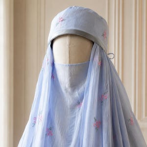 May include: A light blue head covering and face veil set featuring delicate pink floral embroidery scattered across the fabric. The headpiece includes a smooth grey band along the front edge, and the sheer, lightweight material drapes down.