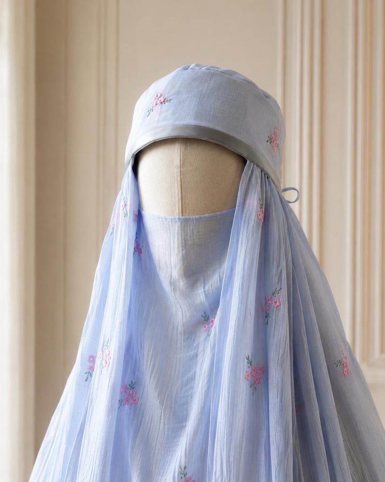 May include: A light blue head covering and face veil set featuring delicate pink floral embroidery scattered across the fabric. The headpiece includes a smooth grey band along the front edge, and the sheer, lightweight material drapes down.
