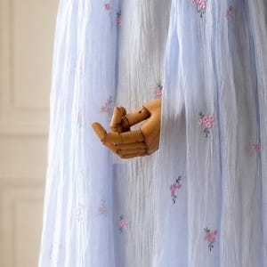 May include: A light blue, semi-sheer, crinkled fabric garment featuring small, scattered pink floral embroidery. The lightweight material has a vertical texture and drapes downward, showcasing delicate needlework patterns across the surface.