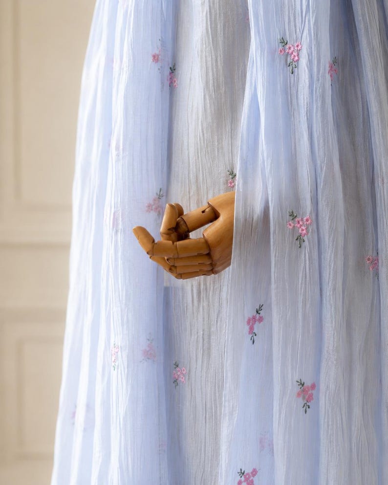 May include: A light blue, semi-sheer, crinkled fabric garment featuring small, scattered pink floral embroidery. The lightweight material has a vertical texture and drapes downward, showcasing delicate needlework patterns across the surface.
