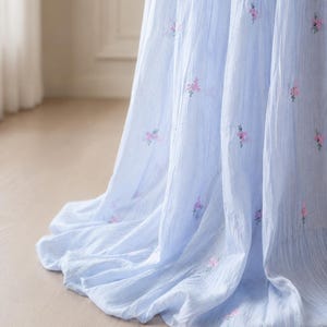 May include: A light blue fabric with a textured, crinkled appearance featuring small, scattered embroidered floral motifs in shades of pink and green. The material drapes in soft, vertical folds, creating a flowing and layered visual effect.