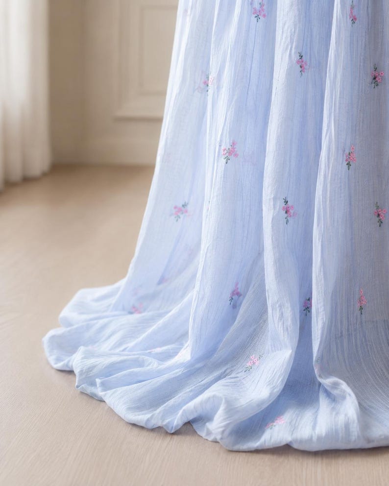 May include: A light blue fabric with a textured, crinkled appearance featuring small, scattered embroidered floral motifs in shades of pink and green. The material drapes in soft, vertical folds, creating a flowing and layered visual effect.