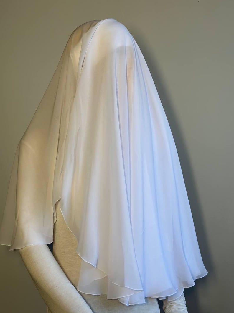 Op de afbeelding: Een witte, doorschijnende sluier gedrapeerd over een mannequin. De sluier is gemaakt van een lichtgewicht stof en heeft een vloeiende, etherische uitstraling.