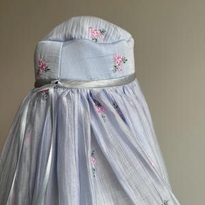 May include: A light blue head covering featuring a structured crown and a flowing, pleated drape. The fabric is adorned with small, embroidered pink floral clusters and green stems. A thin, silver-toned ribbon encircles the base of the crown.