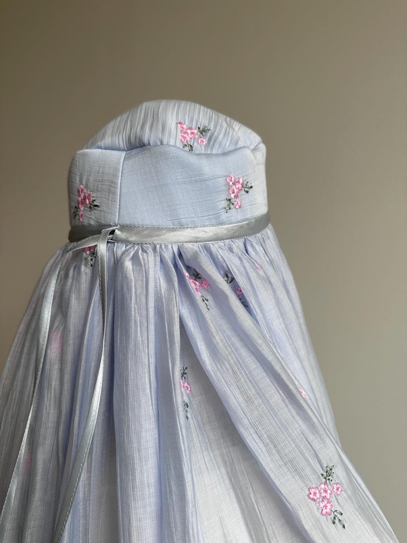 May include: A light blue head covering featuring a structured crown and a flowing, pleated drape. The fabric is adorned with small, embroidered pink floral clusters and green stems. A thin, silver-toned ribbon encircles the base of the crown.