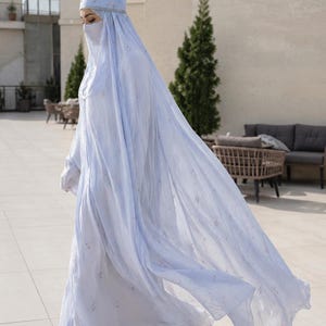 May include: A light blue abaya with a matching head covering and face veil, featuring a delicate floral pattern throughout the fabric. The garment is floor-length with long sleeves and a flowing silhouette, paired with neutral-toned pointed-toe heels.
