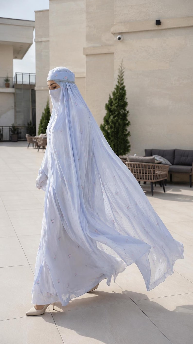 May include: A light blue abaya with a matching head covering and face veil, featuring a delicate floral pattern throughout the fabric. The garment is floor-length with long sleeves and a flowing silhouette, paired with neutral-toned pointed-toe heels.