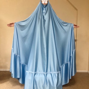 May include: A light blue, flowing, satin abaya with a ruffled hem. The abaya is worn by a person with their face covered.