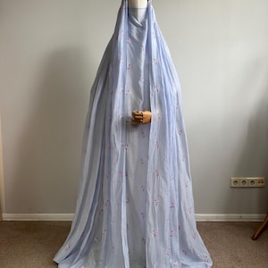 May include: A light blue floor-length abaya with a matching head covering, featuring a delicate pattern of small pink and green floral embroidery scattered across the fabric. The garment has a loose, flowing silhouette and a textured appearance.