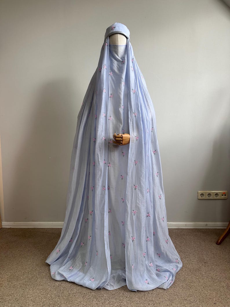 May include: A light blue floor-length abaya with a matching head covering, featuring a delicate pattern of small pink and green floral embroidery scattered across the fabric. The garment has a loose, flowing silhouette and a textured appearance.