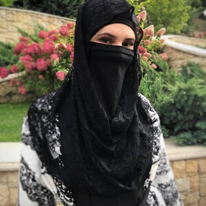 May include: A woman wearing a black lace hijab with a black face covering. The hijab is draped over a white and black patterned dress.