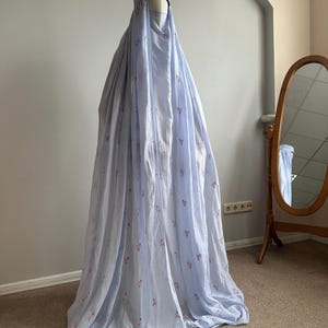 May include: A light blue, floor-length garment featuring a matching head covering and a face veil. The fabric is adorned with a delicate, repeating pattern of small pink floral sprigs, creating a soft, textured appearance on the draped material.