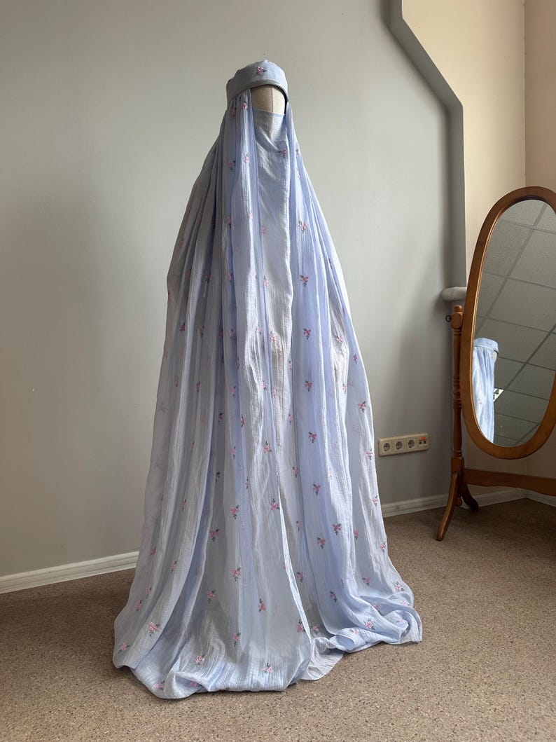 May include: A light blue, floor-length garment featuring a matching head covering and a face veil. The fabric is adorned with a delicate, repeating pattern of small pink floral sprigs, creating a soft, textured appearance on the draped material.
