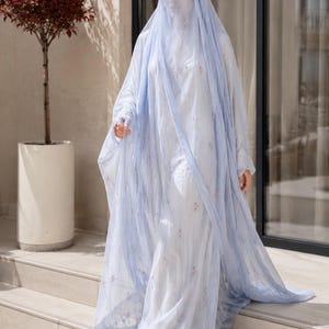 May include: A light blue abaya set featuring a long, flowing outer layer with delicate floral embroidery, paired with a matching head covering and a face veil. The ensemble is crafted from a sheer, lightweight fabric that drapes elegantly.