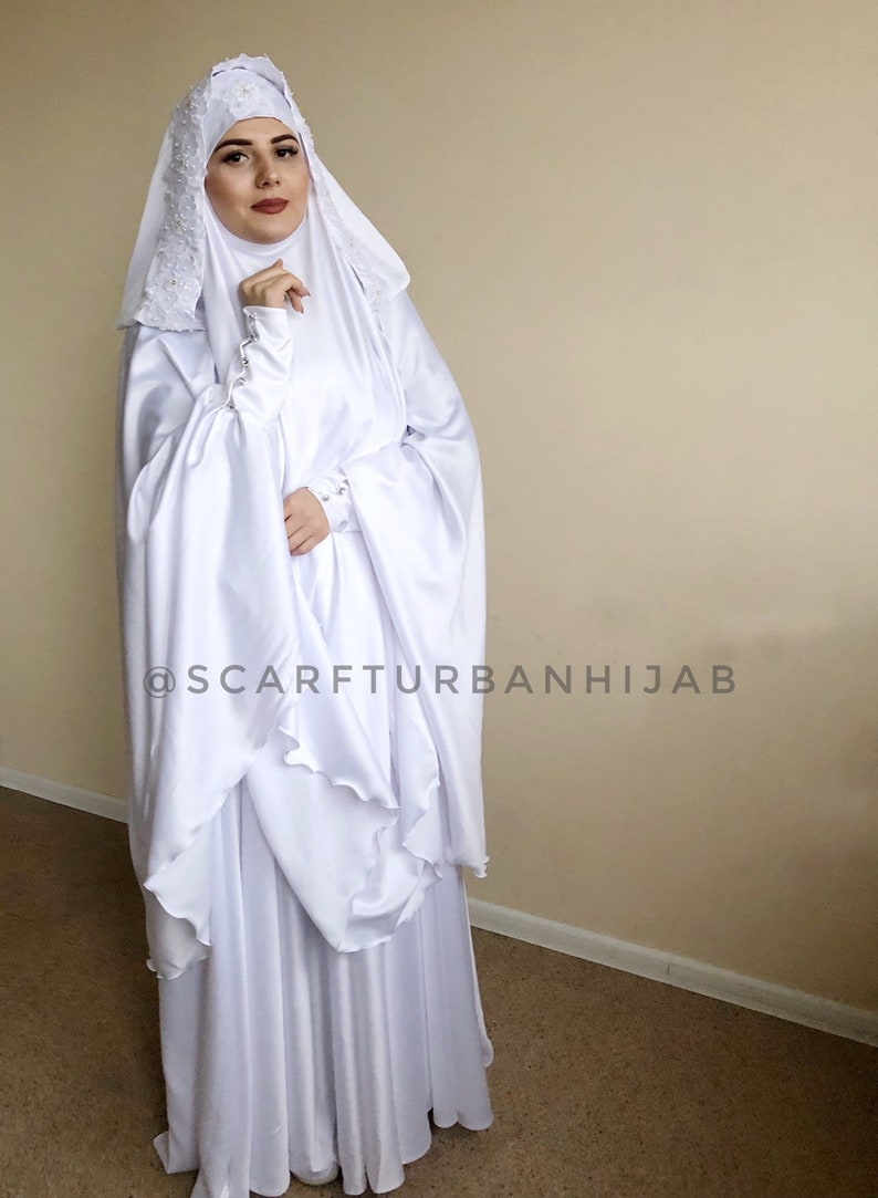 May include: White prayer dress with a long skirt and a matching headscarf. The dress has a ruffled hem and the headscarf has a lace trim. The text "@scarfturbanhijab" is visible on the dress.