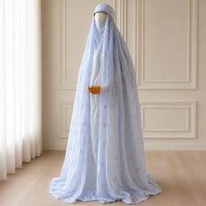 May include: A floor-length, light blue abaya featuring a sheer, flowing outer layer with small, scattered pink floral embroidery. The garment includes a matching head covering and a face veil, creating a cohesive, layered ensemble that drapes to the floor.