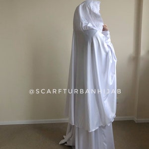 May include: White hooded prayer shawl with lace trim. The shawl is made of a soft, flowing fabric and is designed for prayer. The text "@scarfturbanhijab" is visible on the shawl.