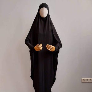 Può includere: Un abaya nero, un capo lungo, esposto su un manichino. L'abaya presenta maniche lunghe e un copricapo. Le mani del manichino sono visibili, posizionate vicino alla parte anteriore del capo. Lo sfondo è un muro grigio chiaro.