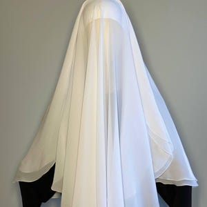 Op de afbeelding: Een witte, doorschijnende stof gedrapeerd over het hoofd en de schouders van een persoon, die het gezicht bedekt. De stof is bij de nek gebundeld en valt in zachte plooien.