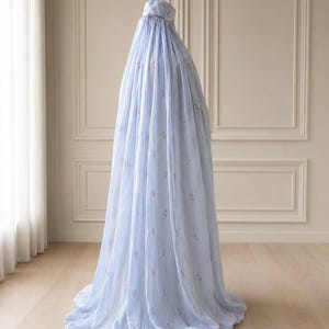 May include: A light blue, floor-length prayer garment featuring a matching head covering and a subtle, repeating pattern of small pink floral motifs. The fabric is lightweight and drapes fully to the floor, creating a voluminous silhouette.
